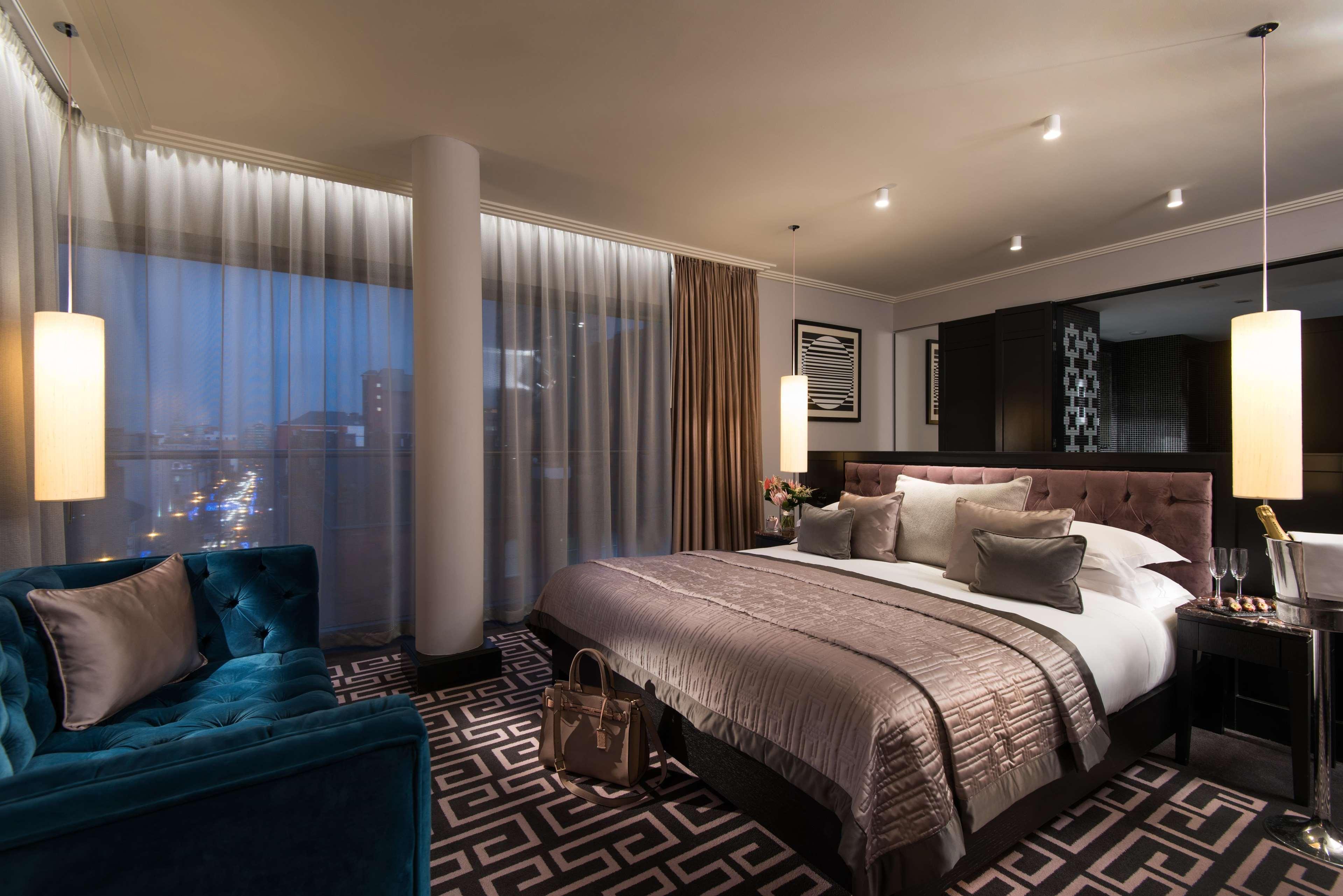 The Fitzwilliam Hotel Belfast Exterior foto The photo shows a luxurious hotel room with a modern design. It features a large bed with decorative pillows and a stylish bedspread, complemented by bedside lamps. To one side, there is a plush turquoise sofa and a coffee table. The room has floor-t
